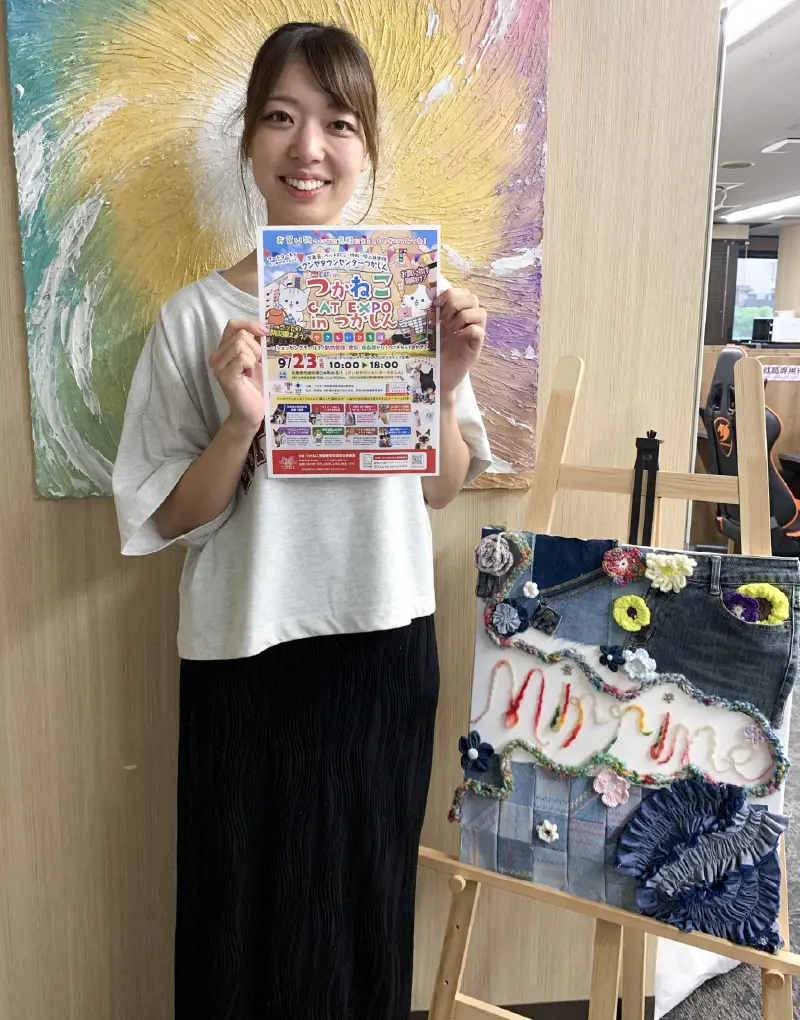 つかねこ CAT EXPO 2025 in つかしんのポスターを持つ女性。背景にはカラフルなアート作品が広がり、会場の華やかな雰囲気を先取りできるシーン。